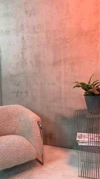 Sillón beige curvado con patas de metal junto a una mesa auxiliar de estructura metálica que sostiene revistas y una planta en maceta contra una pared texturizada.