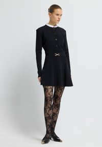 Černé šaty s dlouhými rukávy, přiléhavým korzetem a rozšířenou sukní, se kapsou a zlatým páskem. vzorované černé punčocháče, špičaté boty.