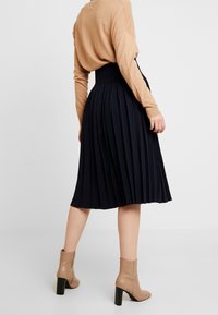 Gonna midi plissettata blu navy con vita alta, abbinata a un top beige a maniche lunghe e stivaletti beige con tacco a blocco.