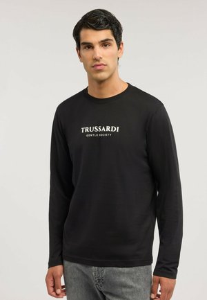 Hombre con cabello oscuro que lleva una camisa negra de manga larga con el texto "TRUSSARDI GENTLE SOCIETY", de pie frente a un fondo liso y claro.