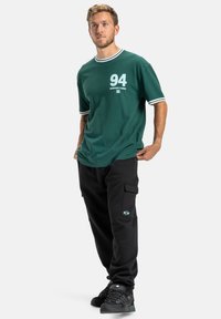 Grünes kurzärmliges Hemd mit weißem Kragen und Logo; kombiniert mit schwarzen Cargo-Hosen mit Taschen; schwarze Schuhe mit strukturiertem Design.