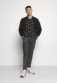Zwarte bomberjack over een geprinte zwarte shirt met rode en witte details, gecombineerd met grijs geruite broeken en witte sneakers met rode accenten.