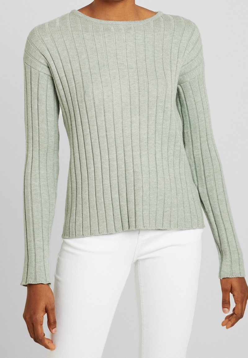 Personne portant un pull côtelé à manches longues vert clair associé à un pantalon blanc sur un fond uni.