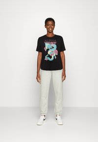 Must mustaga, millel on värviline draakonigraafika ja tekst "ALPHA INDUSTRIES". Kombineeritud heledate hallide dressipükstega ja valgete tossudega.