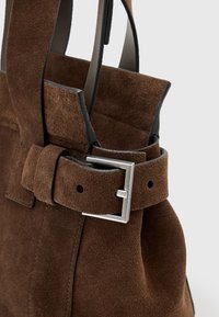 Borsa in suede marrone con ampia tracolla e dettaglio della fibbia rettangolare argentata su uno sfondo neutro.