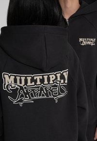 Schwarzer Hoodie mit einem großen grafischen Logo auf dem Rücken in Beige und Weiß. Das Design beinhaltet fetten, stilisierten Text und eine glatte Textur.