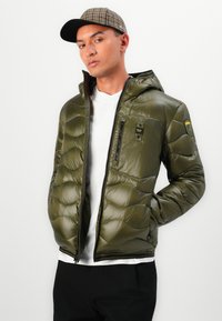 Doudoune vert olive avec capuche, ourlet côtelé, poches zippées et patch logo sur la manche. Portée par-dessus une chemise blanche.