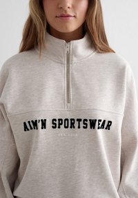 Sudadera gris claro con cremallera de cuarto, con texto bordado "AIM'N SPORTSWEAR" en negro, textura suave y ajuste relajado.