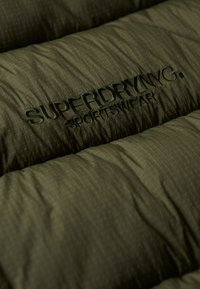Chaqueta acolchada de color verde oliva con textura acolchada y sutil patrón en cuadrícula. Presenta un logotipo bordado de "SUPERDRY" en hilo verde a juego.