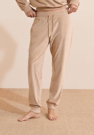 Persoon draagt beige geribbelde joggingbroek met elastische tailleband en trekkers, staat op blote voeten op een gestructureerde beige vloer.