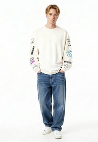 Svetrová mikina krémovej farby so grafickými potlačami na rukávoch, spárovaná s voľnými modrými džínami a bielymi teniskami. Neformálny, uvoľnený strih.