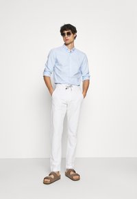 Hellblaues Button-Up-Hemd, weiße Kordelhosen und braune Sandalen. Das Modell posiert mit den Händen in den Taschen vor einem schlichten Hintergrund.