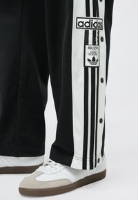 Čierne teplákové nohavice s bielymi bočnými panelmi s tromi čiernymi pruhmi a gombíkmi. Viditeľné sú nášivky s logom Adidas. Biele tenisky so sivými akcentmi.