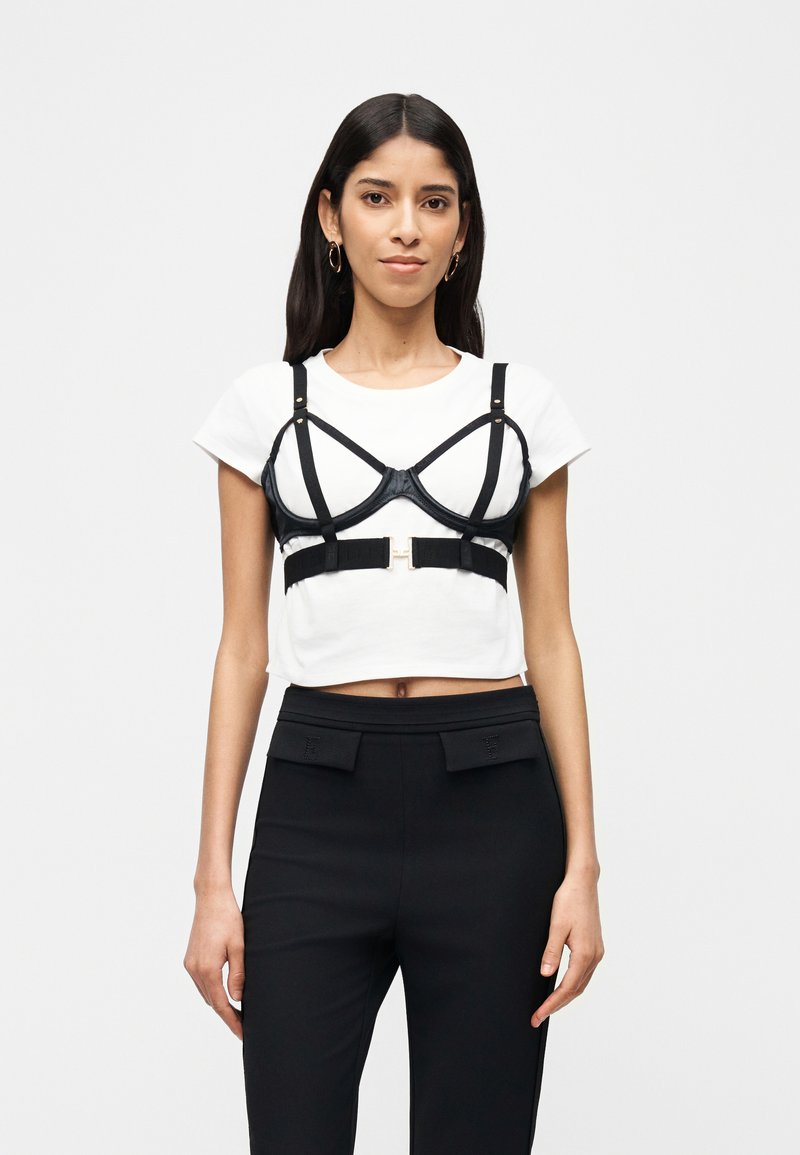 Γυναίκα που φοράει λευκό cropped t-shirt με μαύρο έτοιμο ζώνη από πάνω και μαύρο παντελόνι με ψηλή μέση, σε απλό φόντο.