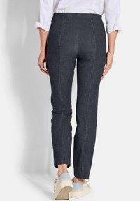 Persoon in donkergrijze strakke broek, witte opgerolde shirt en crèmekleurige sneakers met lichtblauwe accenten, staande tegen een effen achtergrond.