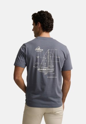 Hombre de espaldas que lleva una camiseta azul grisácea con un dibujo esquemático blanco y etiquetas de un velero en la espalda.