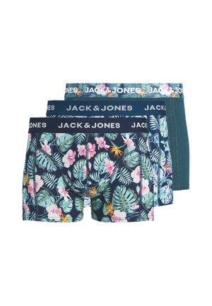 Jonge man met krullend haar, gekleed in blauwe boxershorts met bloemenpatroon en een tailleband met het logo van Jack & Jones, staand tegen een effen lichte achtergrond.