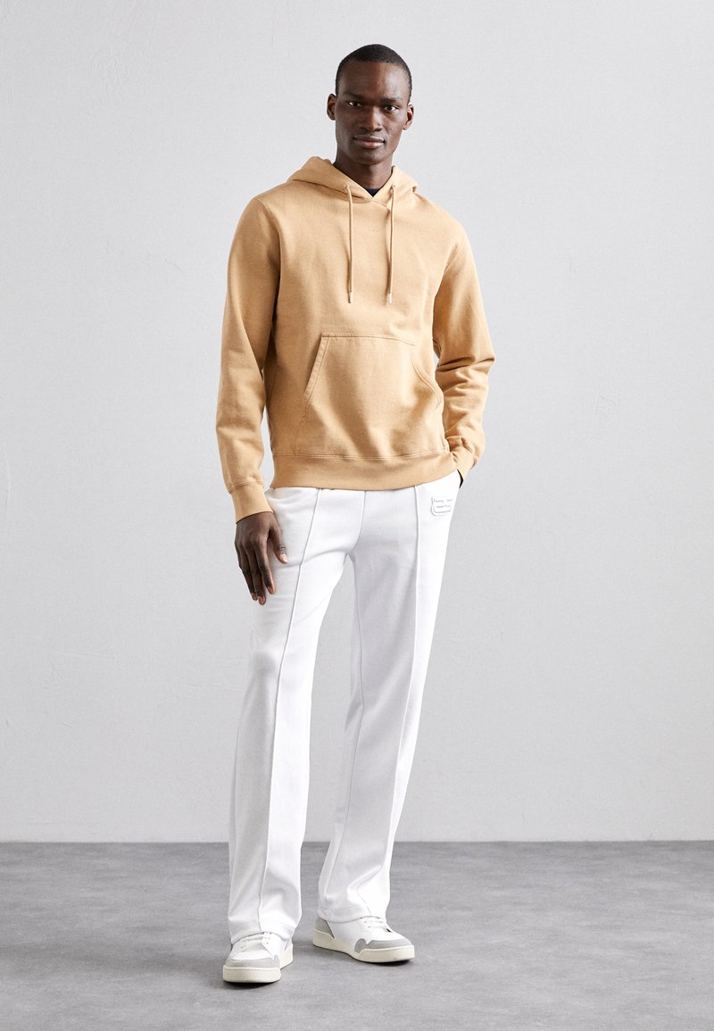 Hoodie din polar bej cu buzunar frontal, asortat cu pantaloni albi cu cusături laterale. Modelul poartă adidași albi. Design minimalist.