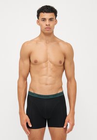 Schwarze Boxer-Brief mit einem grünen elastischen Bund, der mit dem Branding "BJÖRN BORG" versehen ist. Glattes Material, taillierte Passform und bis zur Mitte des Oberschenkels.