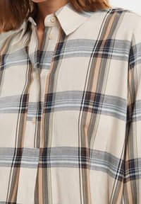Camicia a quadri beige con strisce blu chiaro, marrone scuro e bianche, dotata di colletto classico e chiusura con bottoni. Dettaglio del tessuto texture visibile.