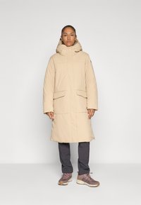 Person iført en beige lang vatjakke med hætte, mørke grå bukser og lyserøde vandresko, stående imod en ensfarvet lys baggrund.