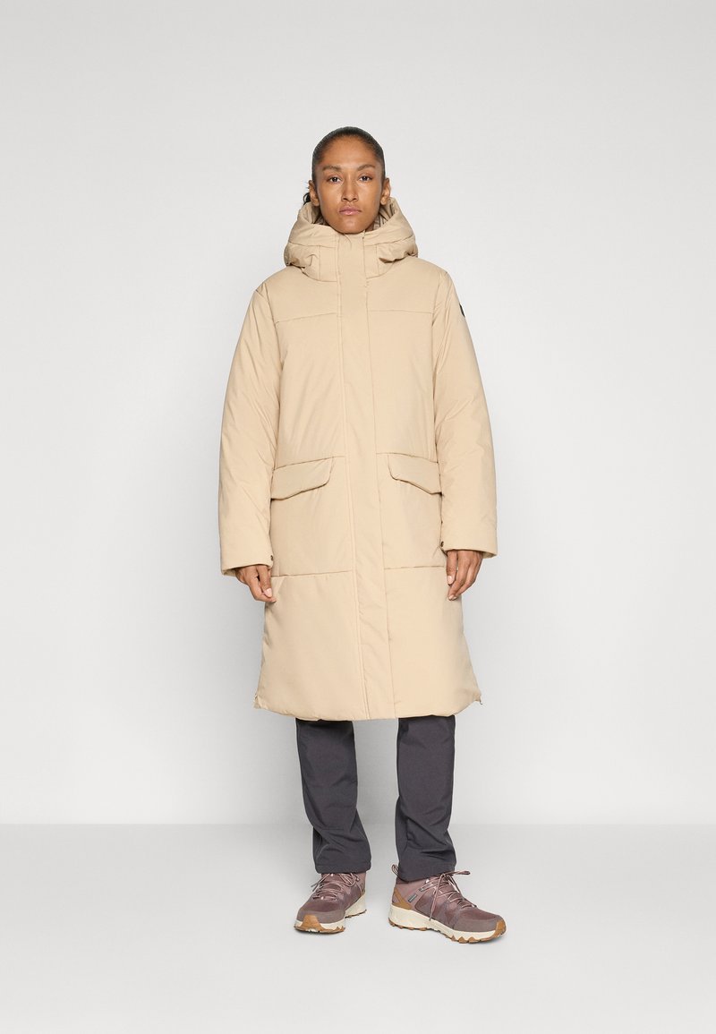 Person iført en beige lang vatjakke med hætte, mørke grå bukser og lyserøde vandresko, stående imod en ensfarvet lys baggrund.