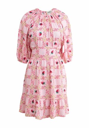 Frau in einem rosa Kleid mit Blumenmuster und Sandalen steht mit gekreuzten Beinen unter einer passenden gebogenen Blumendekoration mit transparenten Vorhängen.
