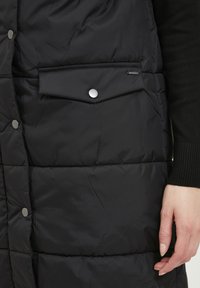 Gilet imbottito nero con un design trapuntato, bottoni a scatto e una tasca frontale con patta. Il materiale appare liscio con una leggera lucentezza.