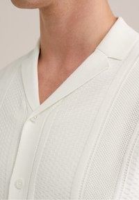 Vit stickad kofta med texturerat mönster, sjalkrage och tre knappar, som visar ett lätt och luftigt tyg.