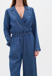 Denim kombinezons ar V veida kakla izgriezumu, garām piedurknēm, elastīgu jostasvietu un sānu kabatām. Audums ir vidēji zils mazgājums ar redzamām šuvēm.