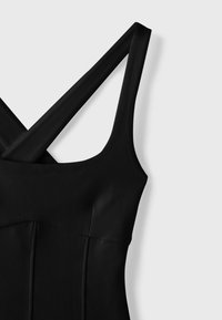 Robe noire sans manches avec de larges bretelles croisées et un corsage ajusté sur un fond clair.