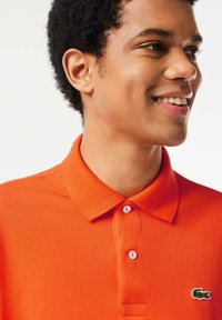 Camisa polo naranja con cuello clásico, tres botones y un patrón texturizado. Un pequeño logo de cocodrilo verde está en el lado izquierdo del pecho.