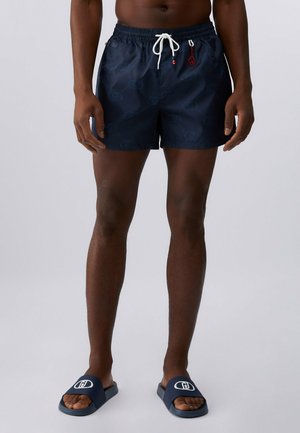 Uomo che indossa pantaloncini da bagno blu scuro con motivo, con coulisse bianca e sandali blu abbinati con logo bianco, in piedi su uno sfondo uniforme.