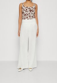 Hedvábný top s květinovým potiskem v béžové a hnědé barvě v kombinaci s bílými kalhotami s širokými nohavicemi. Oblečení má smockovaný pas a bílé špičaté boty.