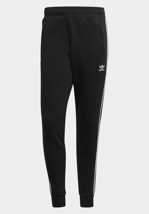 Person iført sorte Adidas træningsbukser med hvide tre striber og logo, parret med hvide Adidas sneakers med sorte striber.