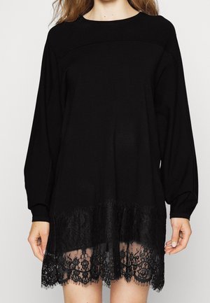 Femme portant une robe noire à manches longues avec un bord en dentelle à l'ourlet, debout devant un fond clair uni.