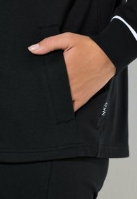 Schwarzes Sportoberteil mit seitlicher Tasche, gerippten Bündchen und einem markanten weißen Akzentstreifen. Weiches Baumwollmaterial mit lockerem Schnitt.