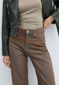 Žena ve hnědých kalhotách s vysokým pasem, béžovém topu a černé kožené bundě, pózující s jednou rukou v kapse a druhou položenou vedle.