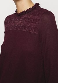 Top lungo bordeaux con maniche lunghe e yoke in pizzo, caratterizzato da un colletto con volant e tessuto texture. La parte inferiore liscia completa la parte superiore dettagliata.