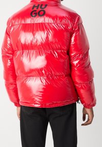 Rote Steppjacke mit glänzender Textur, mit horizontaler Steppung und einem schwarzen "HUGO"-Logo auf dem Rücken, gestaltet mit hohem Kragen und elastischen Bündchen.