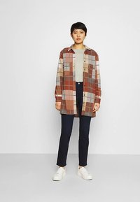 Károvaná košile s knoflíky v teplých tónech, s pohodlným střihem, dvěma náprsními kapsami a zaobleným lemem, nošená s tmavými kalhotami a bílými botami.