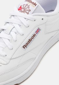 Zapatilla de cuero blanco con suela de goma, que cuenta con la marca Reebok y un emblema de la bandera británica, parte superior texturizada y cordones planos.