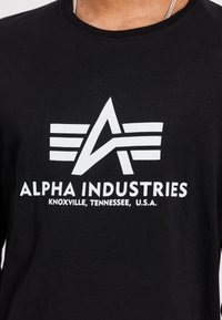 Svart langermet skjorte med et hvitt grafisk logo med vinger og tekst som leser "ALPHA INDUSTRIES, KNOXVILLE, TENNESSEE, U.S.A."
