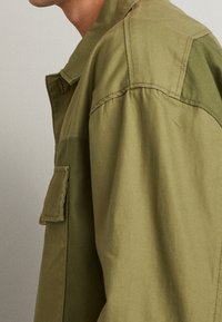 Giacca verde oliva realizzata in tessuto misto cotone, con tasca a pattina sul lato sinistro del petto, vestibilità comoda e dettagli di cucitura minimi.