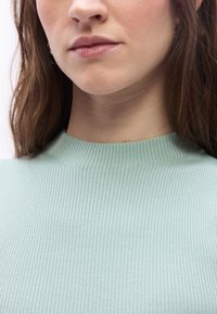 Κοντινό πλάνο στο κάτω μέρος του προσώπου και το λαιμό μιας γυναίκας με μακριά καστανά μαλλιά που φοράει ένα ανοιχτό πράσινο ρ ιμπ πουλόβερ με στρογγυλή λαιμόκοψη.