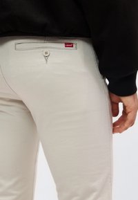 Κοντινό πλάνο ατόμου που φοράει μπεζ παντελόνι Levi’s με κουμπωμένη πίσω τσέπη και μαύρο τοπ, το χέρι ακουμπάει στο πλάι.