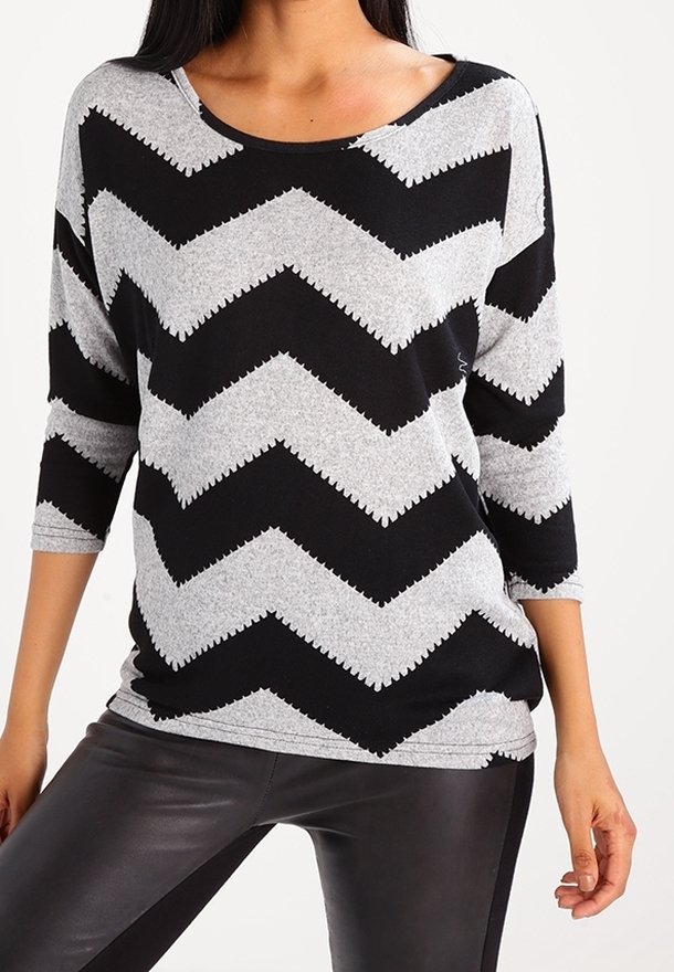 Femme portant un haut à manches longues gris et noir à motif zigzag et un pantalon en cuir noir, debout devant un fond clair uni.