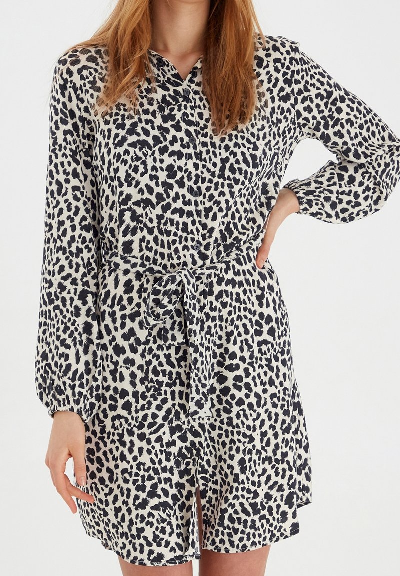 Femme portant une robe à manches longues imprimé léopard noir et blanc, avec une ceinture nouée à la taille et des boutons à l'avant.