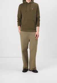 Sudadera con capucha de color verde oliva con un bolsillo frontal y cordones, combinada con pantalones beige claro y zapatos negros con suela gruesa. Detalle de logo minimalista.