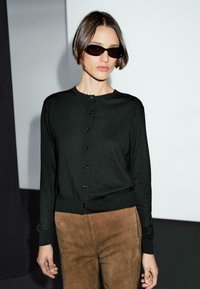 A model poses in a black cardigan and brown suede pants, wearing dark sunglasses against a minimalist background.